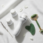 Lifestyle — white facial cleansing brush with replacement heads arranged on white linen beside jade roller and rose quartz gua sha — premium skincare flatlay editorial for visual context only