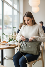 Woman at café accessing front zippered pocket of MultiSac Summerville olive green crossbody bag — multiple compartments for everyday organization
