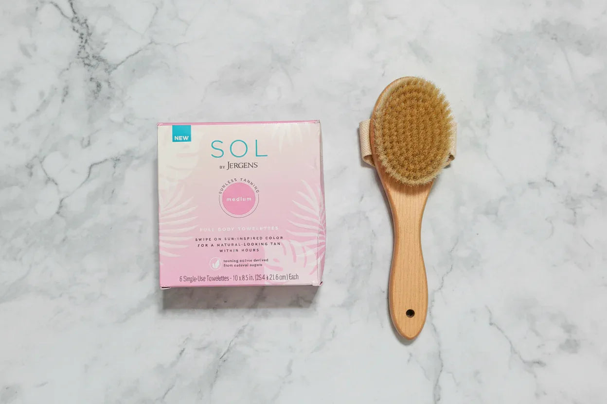 Flat lay top-down view of SOL by Jergens sunless tanning towelettes box beside exfoliating body brush