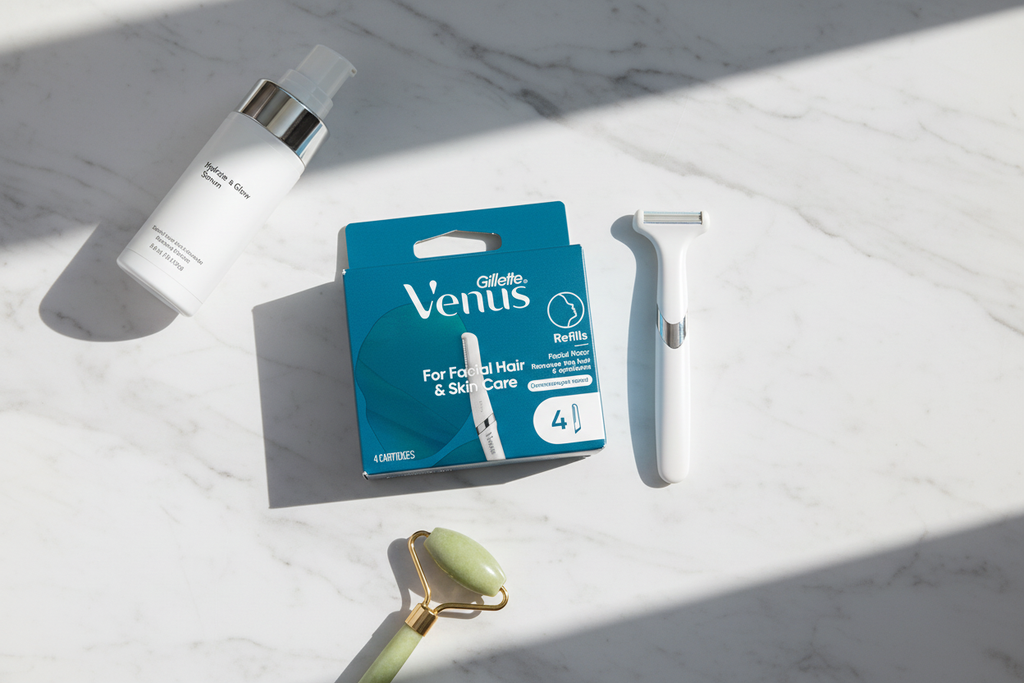 Aesthetic flatlay on white marble with Gillette Venus Facial Refills teal box, slim white razor handle, jade roller, and moisturizer