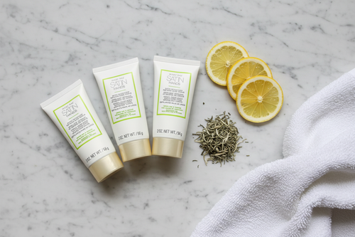 Three white hand scrub tubes arranged next to lemon slices, white tea leaves, and soft towel on marble