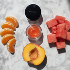 Flat lay of Mary Kay Cheek Glaze Tangerine jar beside fresh mandarin orange slices watermelon chunks and peach on white marble