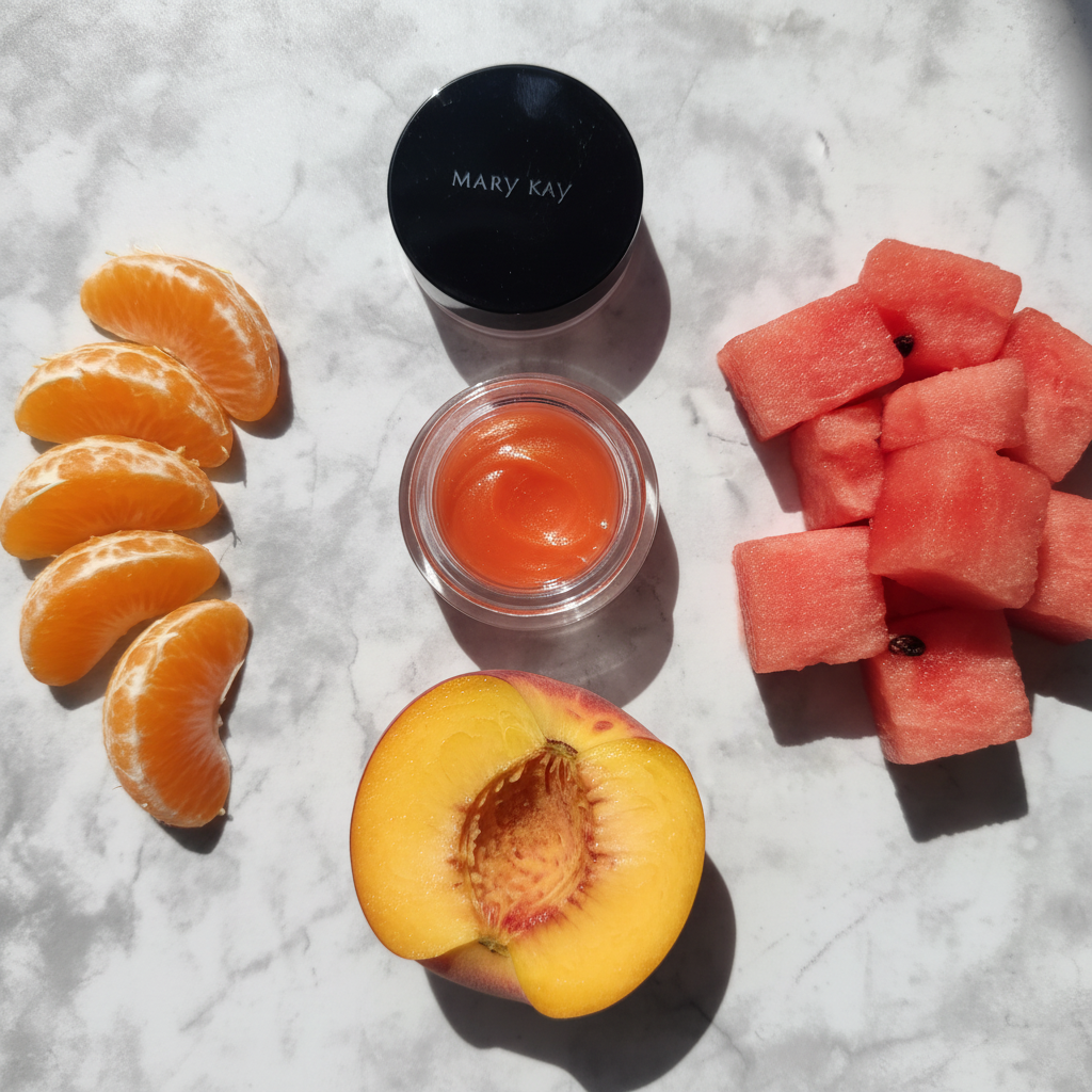 Flat lay of Mary Kay Cheek Glaze Tangerine jar beside fresh mandarin orange slices watermelon chunks and peach on white marble
