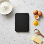 Lifestyle — overhead flatlay of slim black kitchen scale surrounded by flour bowl, eggs, butter, and wooden spoon on white marble — editorial for visual context only