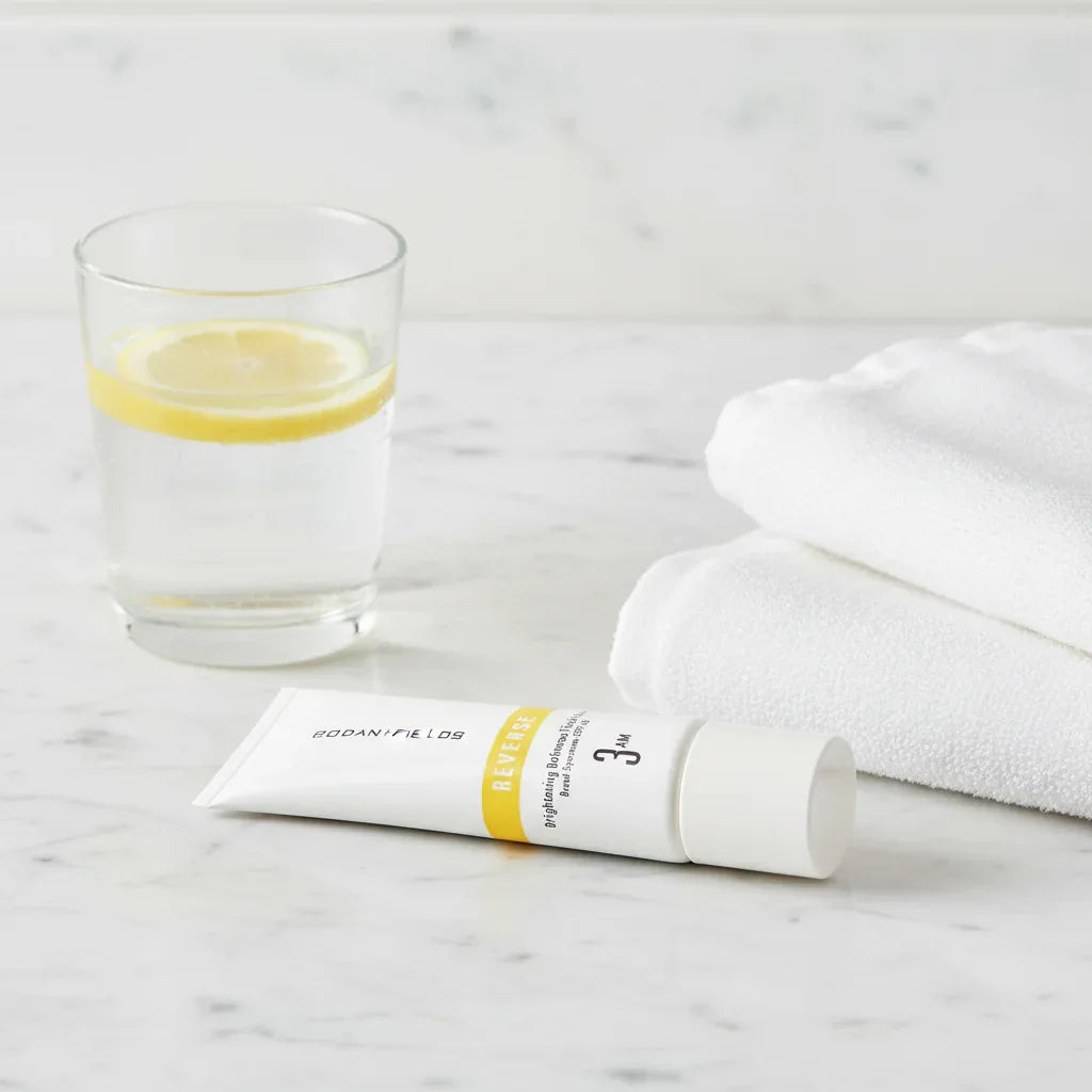 Rodan + Fields Reverse Brightening Defense Fluid SPF 50 tube on contemporary marble bathroom counter next to glass of lemon water and white organic face towel