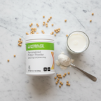 Overhead flat lay of the white Herbalife Nutrition Personalized Protein Powder tub with a tablespoon of white protein powder, a glass of milk, and soybeans on a white marble surface.