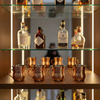 Four hammered copper shot mugs displayed in organized mirrored home bar cabinet with liquor bottles in background