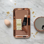 Mary Kay Glowing Finish Illuminating Stick Gold glossy black stick with cream golden shimmer bullet flat-lay on a rose-gold vanity tray with makeup sponge, small mirror, and scattered gold leaf accents on white marble