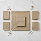 Flat lay of neatly folded natural tan bed sheets and four pillowcases on a white marble surface with sandalwood chips and essential oil vial.