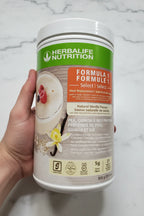 In-hand close-up of Herbalife Formula 1 Select Natural Vanilla protein supplement canister on white marble