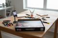 Cupixel x Artsmith Home Decor Set contents laid out on a wooden craft table including wood panels, acrylic paint markers, paint tubes, and device stand with phone showing art instructions.