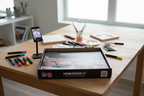 Cupixel x Artsmith Home Decor Set contents laid out on a wooden craft table including wood panels, acrylic paint markers, paint tubes, and device stand with phone showing art instructions.