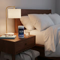 Natural Vitality Calm Mind canister on cozy nightstand with reading lamp, book, and glass of water in peaceful bedroom setting.