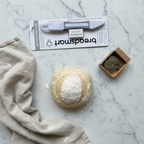Flat lay of artisan bread baking essentials — grey dual-ended bread lame, stainless steel blades, flour dusted linen, and sourdough boule on white marble