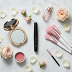 Beauty flat-lay on white marble with sleek black mascara tube pink branding alongside pink makeup brushes compact mirror and flower petals