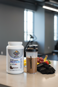 Garden of Life Sport Organic Plant Protein chocolate tub on gym counter with shaker bottle and fresh fruit