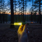 Hand clipping glowing TentBrightz LED stick onto tent rope in forest at twilight.