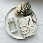 Lifestyle image — clear glass perfume bottle on white vanity tray beside travel journal, dried flowers and pearl necklace, whimsical romantic flatlay for visual context only