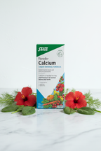 Salus Floradix Calcium Liquid Mineral Formula 250ml retail box on white marble with blue and green botanical props, hibiscus flowers, fennel, and spinach leaves, minimalist studio photography – More4less Outlet
