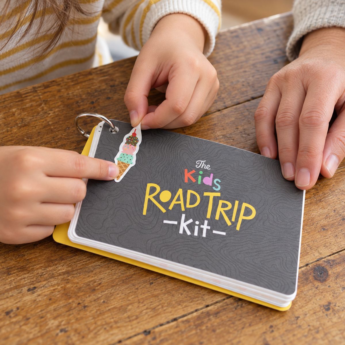 A child applies the included ice-cream sticker to the Inklings Paperie Kids Road Trip Kit cover while an adult steadies it.