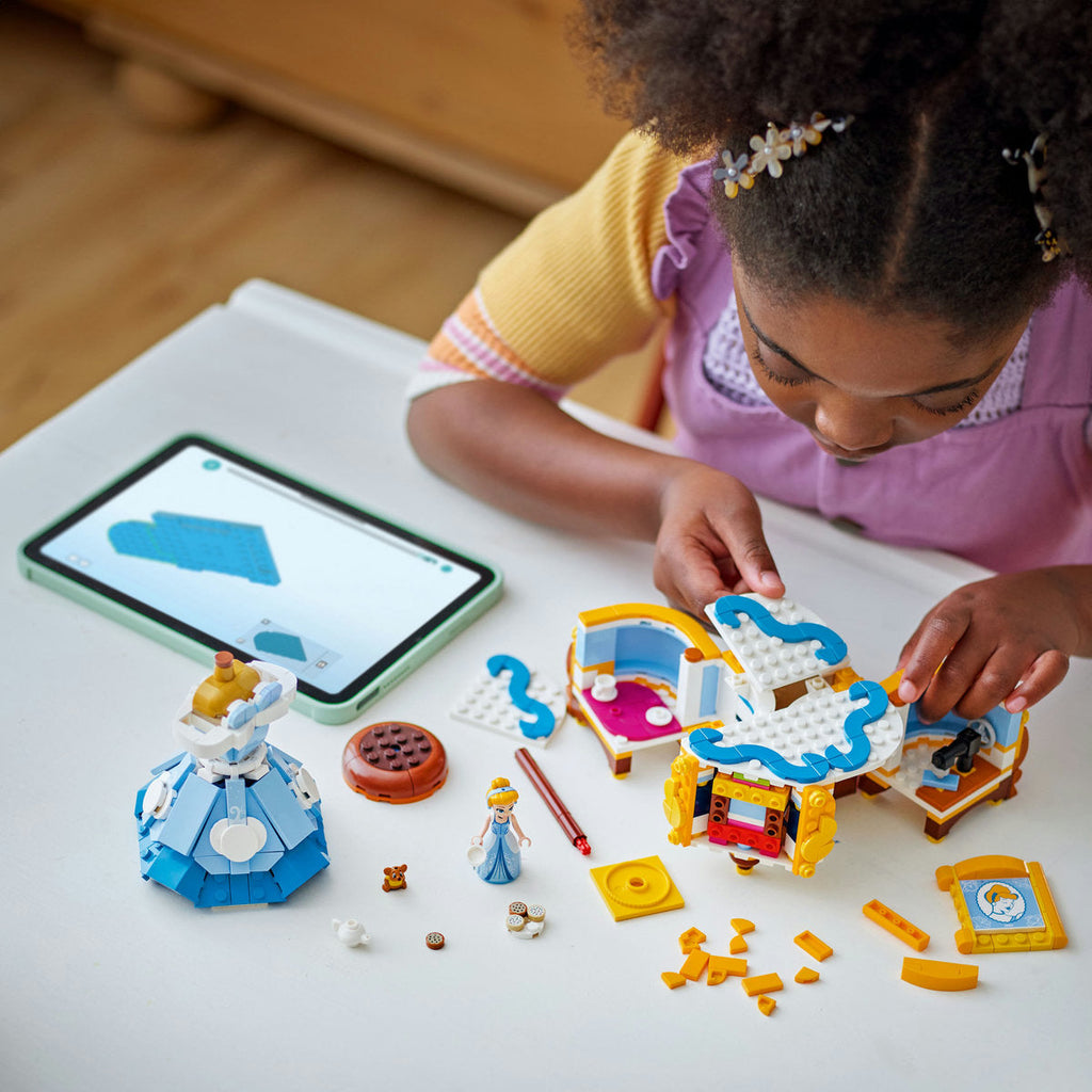 Child assembling LEGO Cinderella’s Dress 43266 beside a tablet, with the dress, display-base pieces and characters on the table