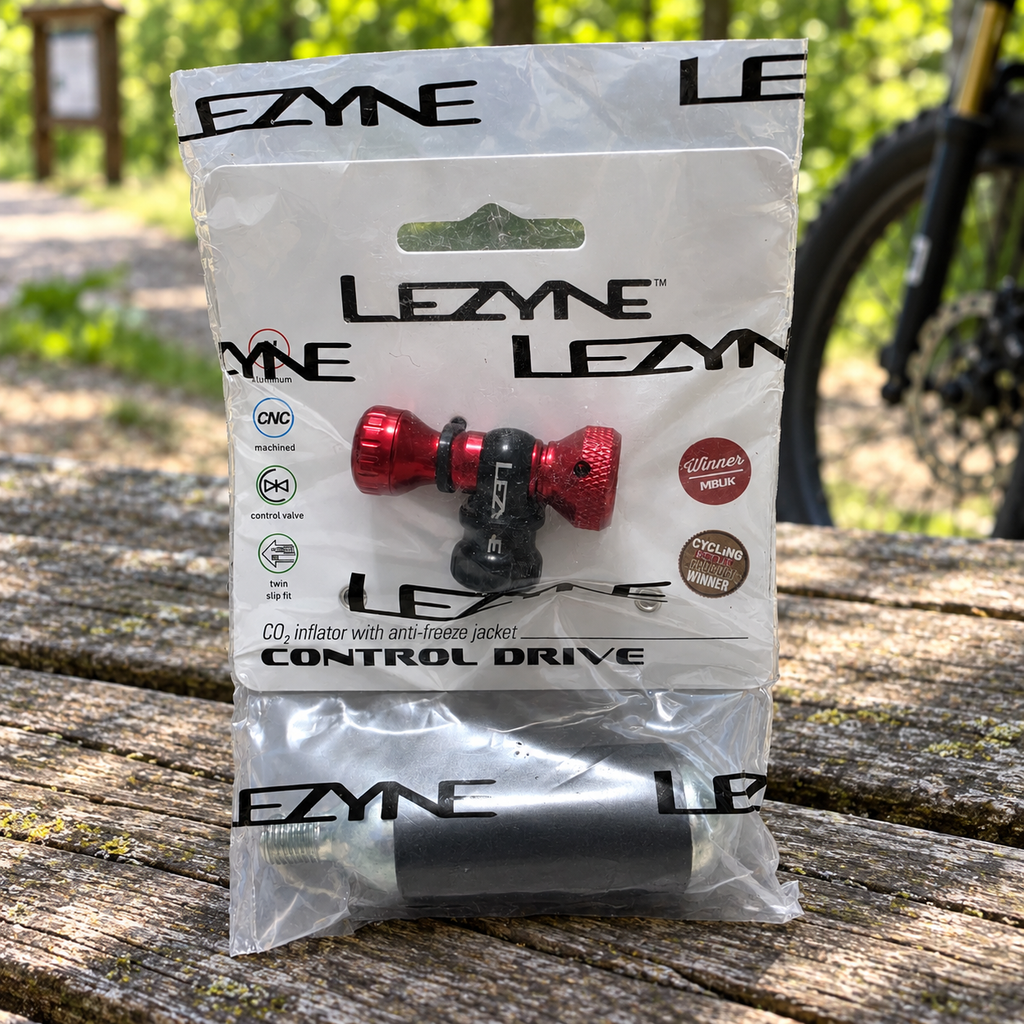 Packaged red Lezyne Control Drive CO2 inflator and 16g cartridge on a picnic table at a cycling trailhead; AI-staged lifestyle composite.