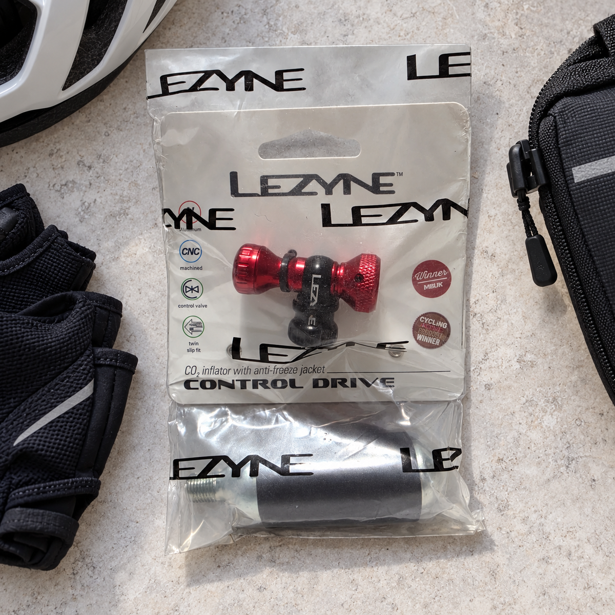 Packaged red Lezyne Control Drive CO2 inflator with 16g cartridge beside a saddlebag and cycling gloves; AI-staged lifestyle composite.