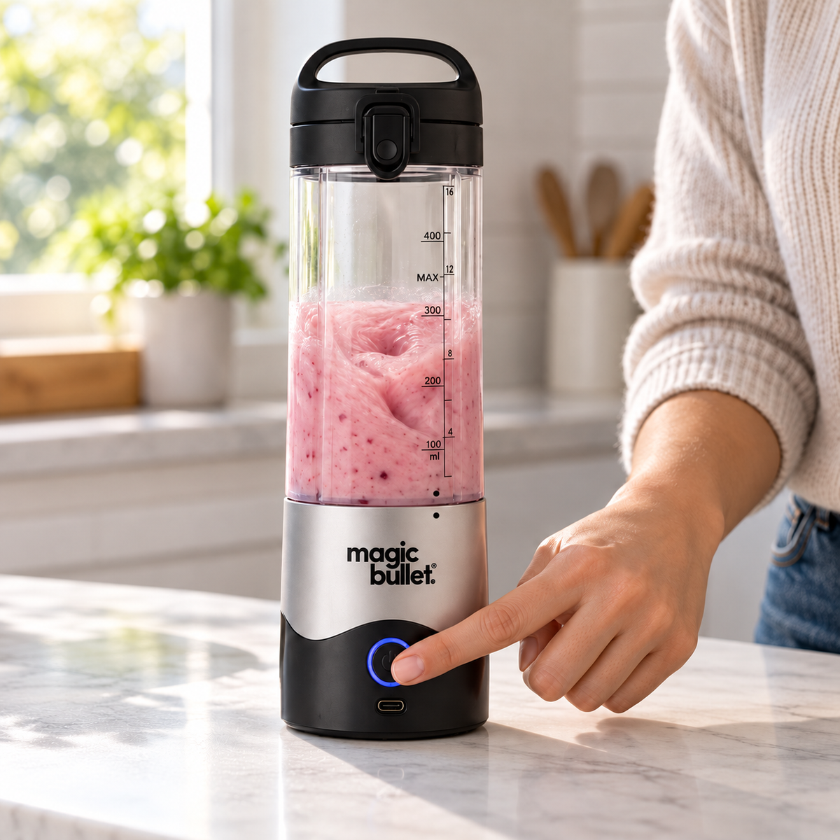 Illustrative lifestyle image of an adult pressing the front power button on a silver Magic Bullet portable blender with its lid locked and smoothie below the MAX line.