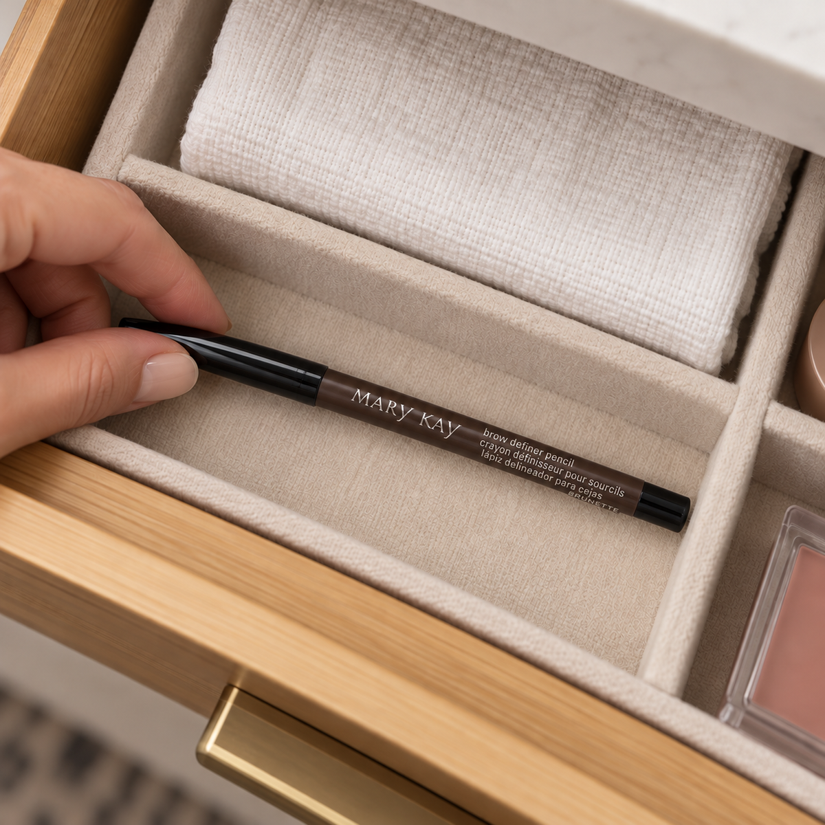 A hand placing a capped Mary Kay Brunette Brow Definer Pencil in a divided makeup drawer.