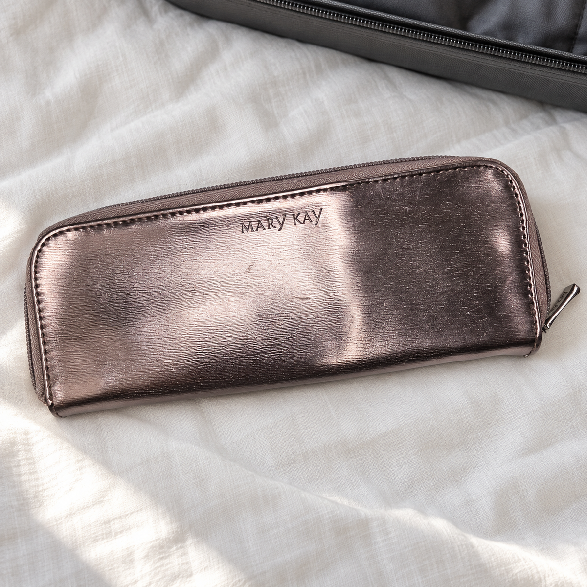 Closed metallic Mary Kay eye brush case on ivory linen beside a suitcase
