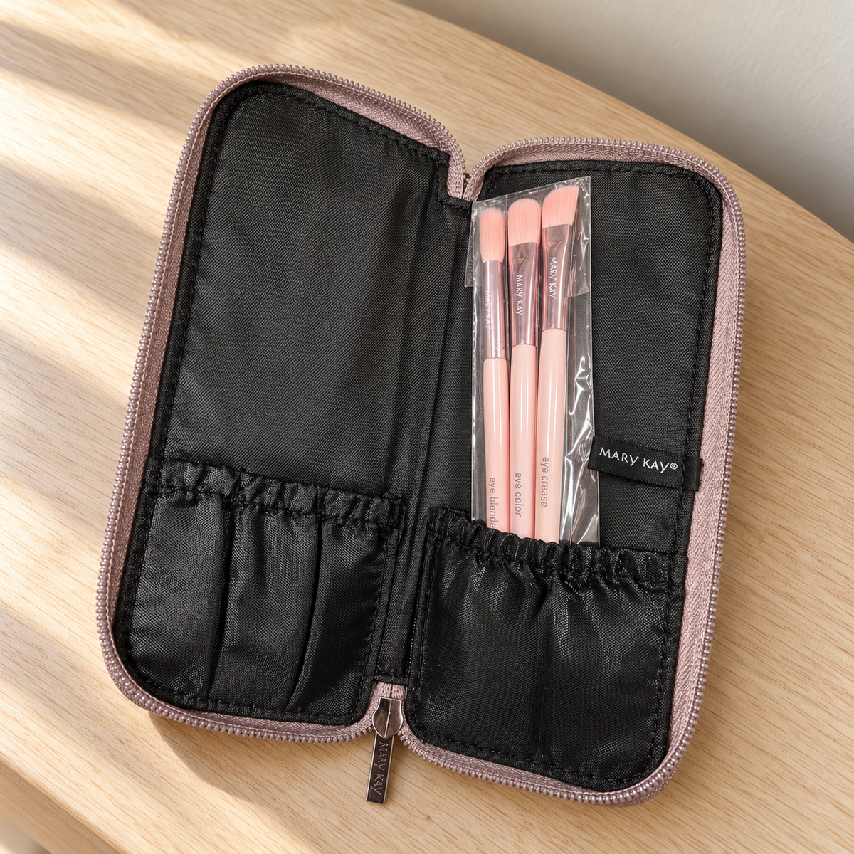 Three pink Mary Kay eye brushes in sleeves inside the open metallic case on an oak vanity