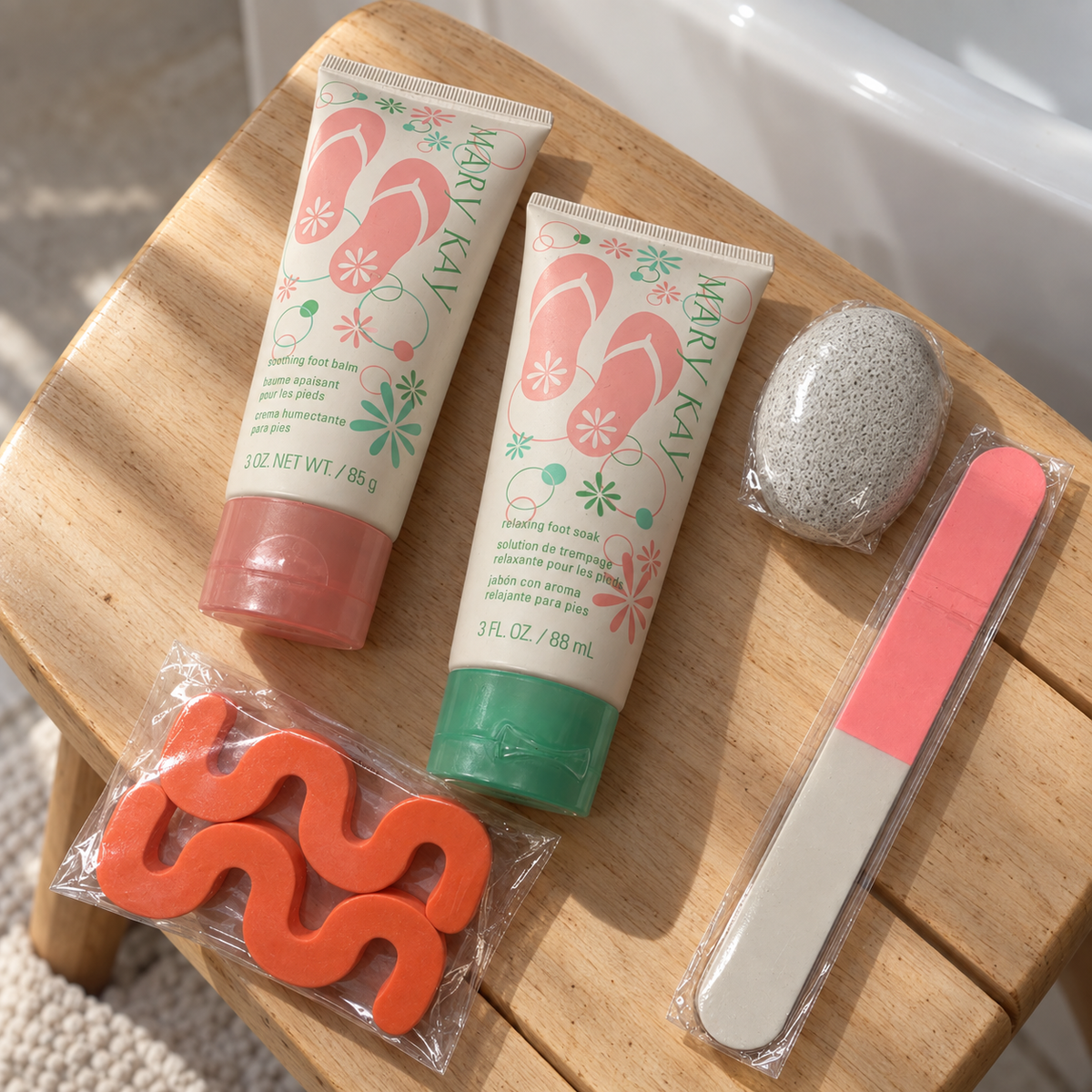 Mary Kay foot balm, foot soak, nail file, two orange toe separators and sealed gray pumice stone arranged on a wooden bath stool