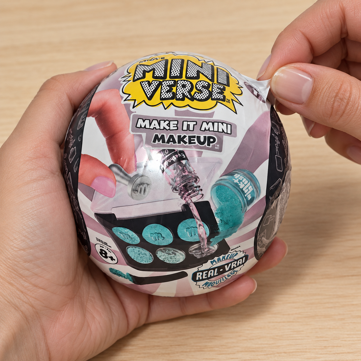 Illustrative adult hands peeling the wrapper from a closed Miniverse Make It Mini Makeup capsule.
