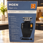 Moen Host EX75c 3/4 HP retail carton in a contemporary kitchen counter setting; package illustration shows the black corded disposal.
