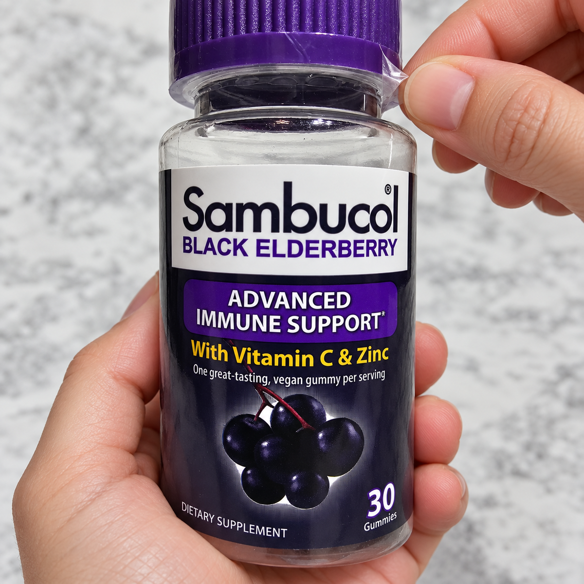 Hands removing the clear cap wrapping from a Sambucol Black Elderberry gummy bottle.