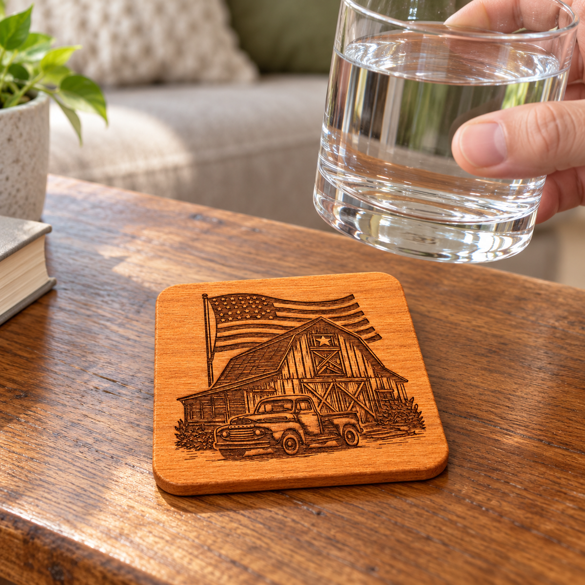 Barn, flag and pickup-truck engraved wood coaster beneath a raised clear drinking glass on a coffee table.