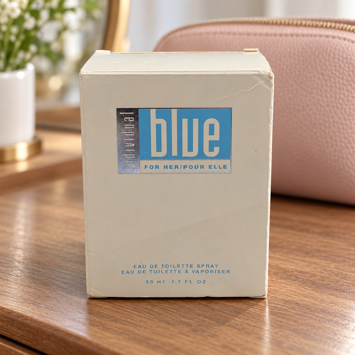 AI-generated context: Avon Individual Blue For Her 50 ml retail carton with visible wear on a wood vanity with a pink pouch; setting props not included.