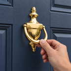 AI-generated context: adult lifting the hinged ring of a Baldwin Colonial brass knocker on a navy door; door is not included.