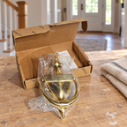 AI-generated context: the bagged Baldwin Colonial brass knocker and opened box on a home renovation table; cloth and setting props are not included.