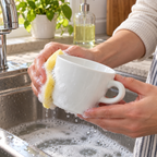 Hands wash a smooth white Bernardaud Opus teacup with a soft sponge at a sink.