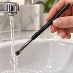 Rinsing the dark bristle tips of a Mary Kay makeup brush while keeping its handle above the water.