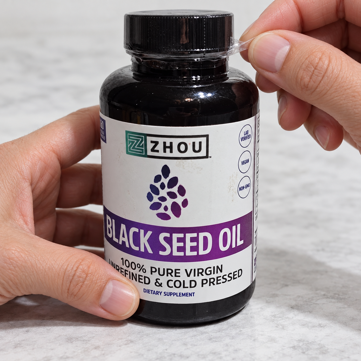 Hands removing the cap wrapping from a Zhou Black Seed Oil bottle.