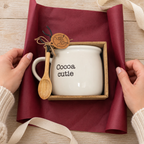 Cocoa cutie mug and wooden spoon in their brown display box as hands fold burgundy gift wrap around the set.