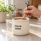 Hand stirring cocoa in the Cocoa cutie mug with the included wooden spoon on a kitchen counter.