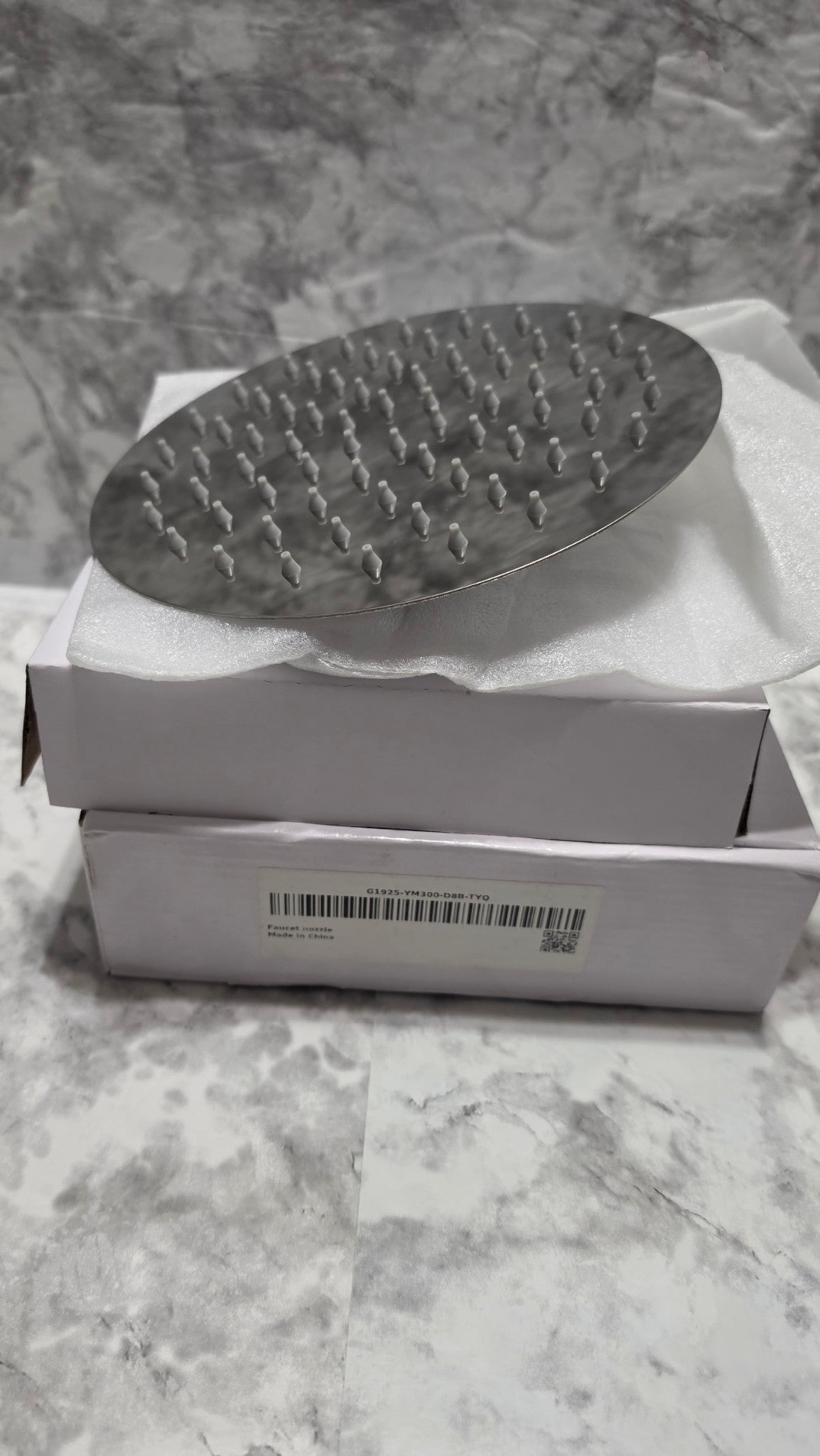 Actual round chrome-look shower head resting on its original foam packaging.