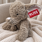 AI-generated lifestyle scene of the gray RUSS elephant plush with white trunk tip and attached red tag on a bedroom chair. Props not included.