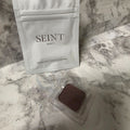 SEINT London eyeshadow pan in a clear protective tray beside its original silver pouch on a marble-pattern background