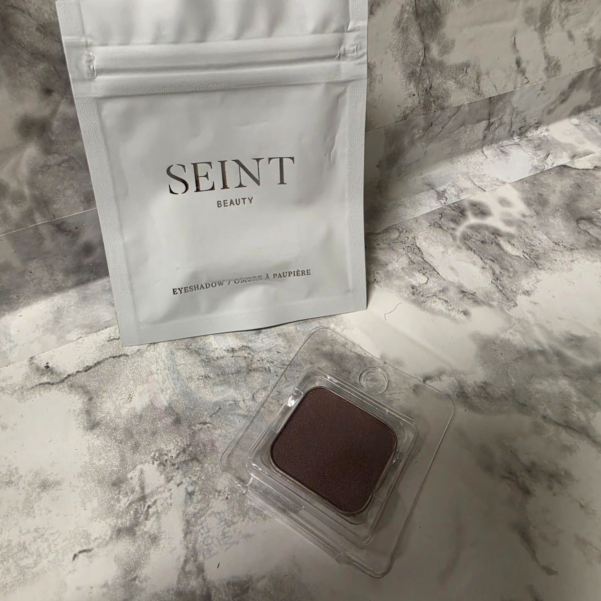 SEINT London eyeshadow pan in a clear protective tray beside its original silver pouch on a marble-pattern background