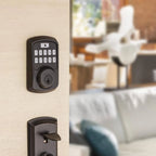 A matte black electronic keypad door lock mounted on a light wood door, with a matching dark handle visible below and a blurred living room interior in the background.