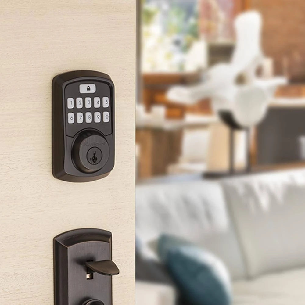 A matte black electronic keypad door lock mounted on a light wood door, with a matching dark handle visible below and a blurred living room interior in the background.